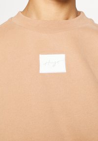 Sudadera color canela con cuello redondo, presenta una textura sutil y un parche blanco rectangular con texto bordado. Diseño clásico para uso casual.
