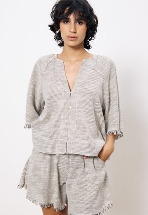 Jeune femme aux cheveux courts et bouclés portant une blouse ample à rayures avec des boutons et un short effiloché assorti, les mains dans les poches.