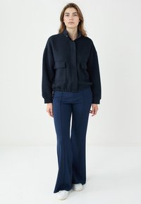 Vrouw staande in een gestructureerde zwarte jas met voorzakken, marineblauwe flared broek en witte sneakers tegen een effen lichte achtergrond.