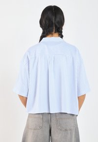 Camicia a maniche corte con colletto, caratterizzata da strisce verticali blu e bianche. Vestibilità ampia con schiena plissettata per dettagli extra.