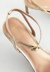 Sandali con tacchi alti a strappi metallici dorati presentano una soletta rosa liscia, un accento circolare dorato sul cinturino e una superficie testurizzata.