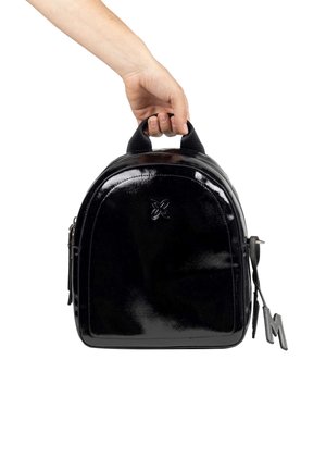 Mochila de charol negro de imitación con forma redondeada, cierre con cremallera, asa superior y un charm decorativo en forma de "M" colgando de un lado.