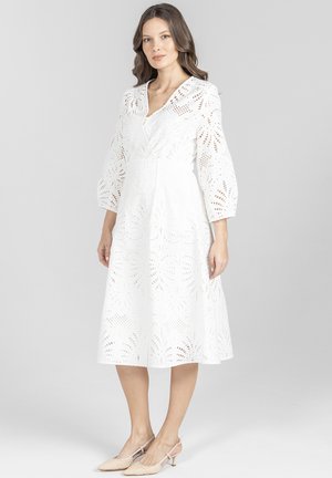 Kobieta ubrana w białą sukienkę midi z ażurowymi wzorami i rękawami 3/4 typu balonowego, w zestawieniu z jasnobeżowymi sandałami na niskim obcasie ze zapięciem na pasek.