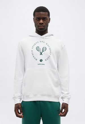 Uomo con felpa bianca con cappuccio con racchette da tennis verdi, grafica di palla e testo "Monaco Sports and Racquet Club", abbinata a pantaloni verdi.