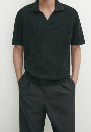 Schwarzes Strick-Polohemd mit V-Ausschnitt und kurzen Ärmeln, strukturierter Stoff, kombiniert mit dunklen Hosen. Beobachtete Hände in den Taschen.