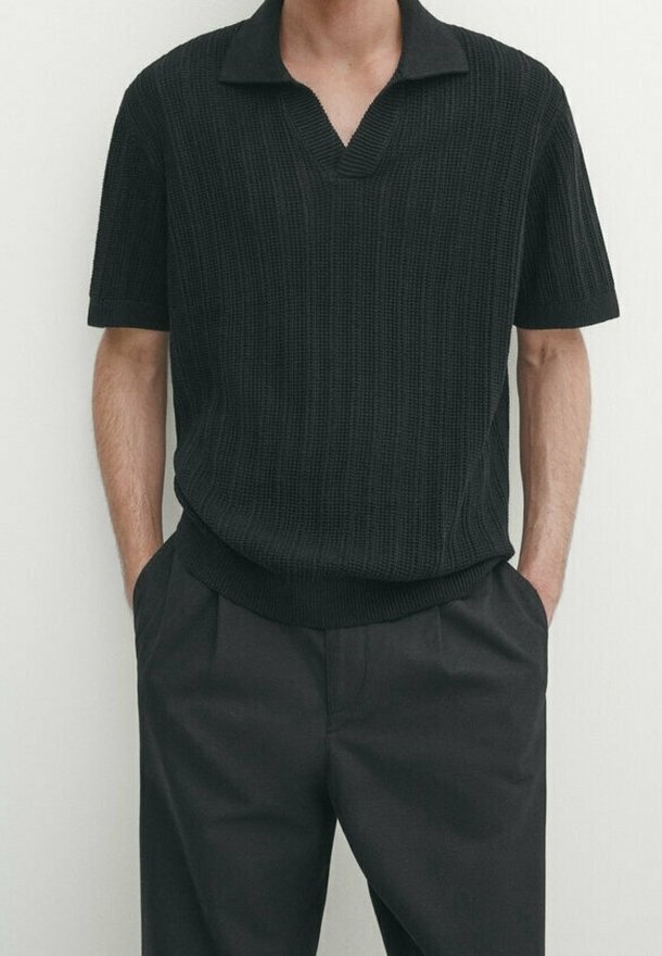 Polo nero in maglia con scollo a V e maniche corte, tessuto texturizzato, abbinato a pantaloni scuri. Mani nelle tasche.