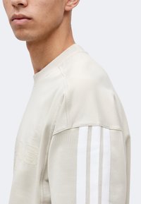 Jeune homme portant une chemise beige à manches longues avec trois bandes verticales blanches sur l'épaule et le haut du bras.