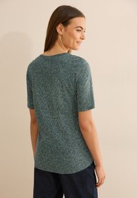 Triko s krátkým rukávem v zajímavé modrozelené barvě s geometrickým vzorem, vyrobené z měkké látky, s kulatým výstřihem a rovně střižené, nošené s tmavými kalhotami.