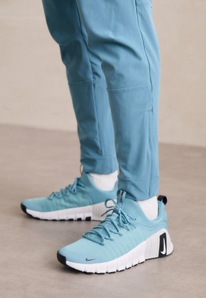Personne portant des chaussures de sport bleu clair avec des semelles blanches et un pantalon bleu clair assorti, debout sur un sol lisse.