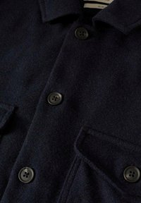 Manteau en laine bleu marine foncé avec quatre boutons noirs et poche à rabat visible sur le devant, montrant le col et un gros plan sur le tissu texturé.