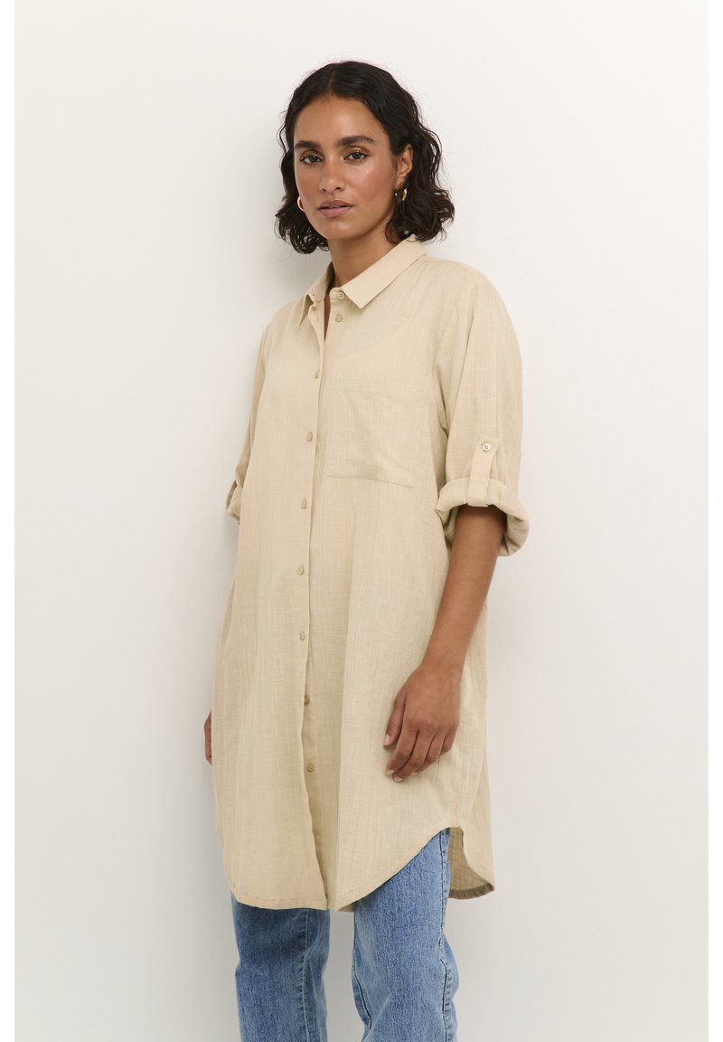 Chemise en lin beige avec col, fermeture à boutons, poche poitrine unique et manches retroussées. La chemise a une silhouette relaxée et allongée.