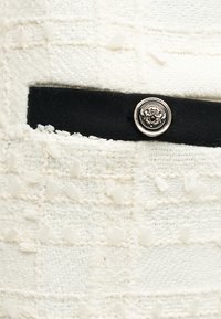 Λευκό υφασμάτινο ύφασμα με μαύρη υφασμάτινη λωρίδα που διαθέτει ένα μικρό ασημένιο κουμπί με ανάγλυφο σχέδιο θυρεού.