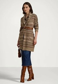 Kvinna klädd i en mönstrad beige kofta med bälte i midjan, blå denimkjol, bruna högklackade stövlar, hållande en brun handväska.