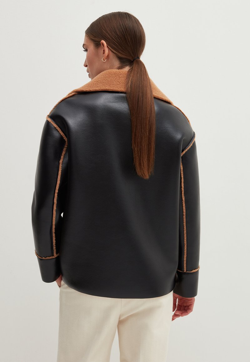 Schwarze Lederjacke mit braunem Fleece-Kragen und -Stichen. Übergroßer Schnitt, glatte Textur, leicht abgerundeter Saum und lange Ärmel.