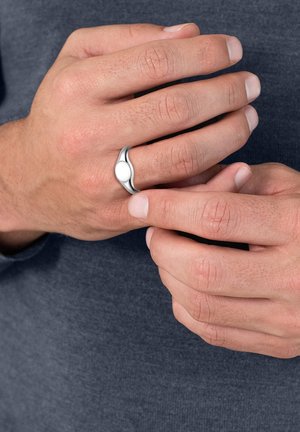 Anello in metallo argento con un design liscio e curvato. Presenta un centro bianco rotondo e una finitura lucida. Le mani sono posate in modo casuale, mentre afferrano.