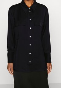 Chemise noire à manches longues avec col à boutons et deux poches poitrine. Tissu lisse, silhouette droite, présentant des boutons et des poignets blancs.