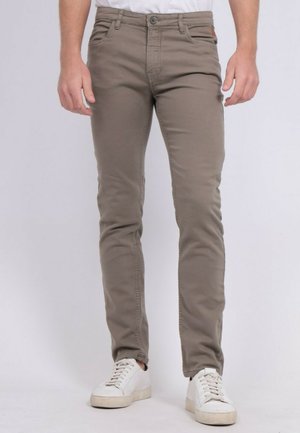 Homme portant un pantalon marron clair ajusté et des baskets blanches, debout devant un fond uni clair.