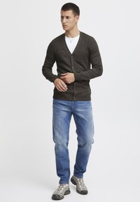 Mørkebrun strikket cardigan med V-hals, knappelukking og ribbestrikket skulderdesign, kombinert med lyseblå jeans og grå sneakers.