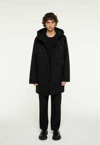 Sort parka med frontlynlås, hætte og to sidelommer. Bæres over et sort outfit og parret med sorte klodsede sko. Minimalistisk design.