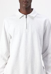 Polo de manga larga gris con cuello de cremallera, fabricado en un tejido suave, con un diseño de cuello y una textura sutil. Etiqueta visible en el lateral.