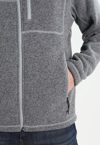 Szara polarowa kurtka z zamkiem z przodu, kieszeniami po bokach i kontrastowymi jasnoszarymi stworzami. Miękka tekstura i luźny krój.