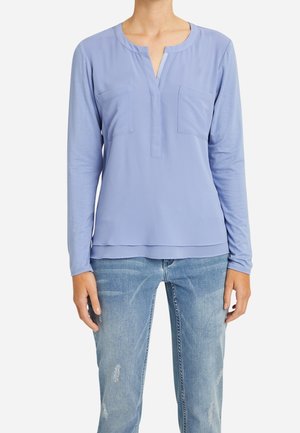 Woman wearing light blue long-sleeve blouse with two chest pockets and faded blue jeans against a white background.