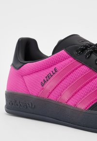Rózsaszín hálós sportcipők fekete szintetikus részletekkel, három fekete csíkkal, kerek orral és texturált gumitalppal. A "Gazelle" logó látható.