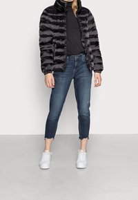 Giacca a vento nera con cuciture orizzontali, colletto alto e polsini elastici, abbinata a jeans cropped in denim scuro e sneakers bianche.