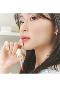 Gloss à lèvres dans un tube crémeux avec un applicateur rouge, tenu par une main aux ongles rose clair. Le produit a l'air brillant et légèrement pigmenté.