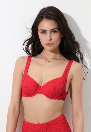Donna con lunghi capelli scuri che indossa un reggiseno rosso con motivo e slip a vita alta abbinati, in piedi davanti a uno sfondo bianco.