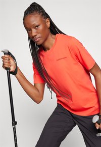 T-shirt orange vif à manches courtes avec un logo sur le côté gauche de la poitrine, associé à un pantalon foncé. La personne tient un bâton de randonnée avec une prise grise.