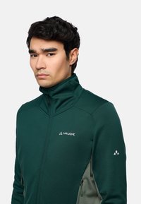 Giacca da uomo verde con zip e colletto alto, texture a rete, dotata di logo Vaude, tasche laterali e accenti grigi a contrasto.