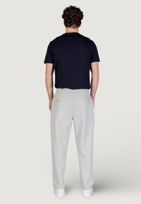 Homme de dos portant un t-shirt noir à manches courtes, un pantalon gris clair avec des poches arrière et des baskets blanches sur un fond uni.