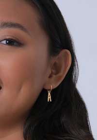 Boucles d'oreilles en or en forme d'anneau avec un charm représentant une lettre 'a' en minuscule, surface texturée et taille moyenne, fixées près du lobe de l'oreille.