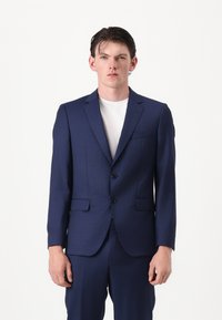 Traje azul marino hecho de tela texturizada, con cierre de dos botones, solapas con muesca y dos bolsillos frontales, combinado con una camiseta blanca.