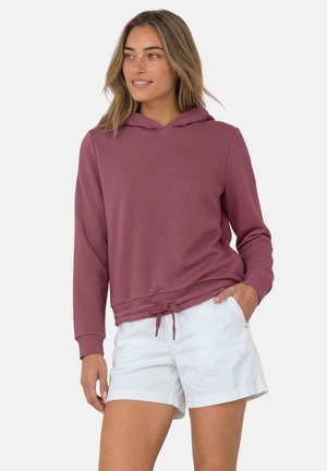 Kvinde med lyst brunt hår iført en lilla hættetrøje og hvide shorts, med den ene hånd i lommen, der kigger til siden mod en ensfarvet baggrund.