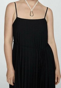 Schwarzes plissiertes Kleid mit dünnen Trägern und tailliertem Schnitt. Ausgestattet mit einem strukturierten Stoff und einer weißen geflochtenen Halskette mit einem Metallelement.