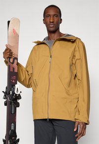 Wodoodporna brązowa kurtka narciarska z zamykanymi na zamek kieszeniami i kapturem, trzymana przez osobę obok nart i wiązań na neutralnym tle.