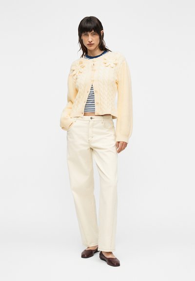 Γυναίκα που φοράει εκρού ζακέτα με σχέδιο πλεξούδας και λεπτομέρειες με λουλούδια, ριγέ πουκάμισο, εκρού παντελόνι ψηλόμεσο και καφέ επίπεδα παπούτσια, στέκεται με το χέρι στην τσέπη.