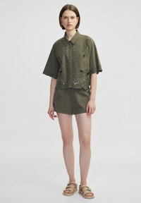 Chemise olive vert à manches courtes avec des détails en dentelle et des accents métalliques, associée à un short assorti. Le tissu semble doux et léger.