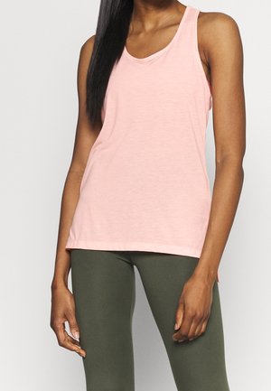 Femme portant un débardeur rose clair sans manches et des leggings vert olive, debout avec les bras détendus contre un fond uni.