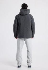 Dark gray padded jacket with a large hood, featuring a smooth texture and minimalistic design. Model wears gray sweatpants and sneakers.