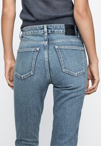 Jeans en denim bleu clair avec taille haute, cinq poches et une étiquette de marque en cuir au dos. Présence de marques et de texture délavée.
