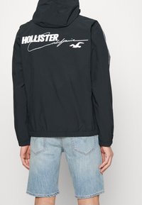 Černá mikina s kapucí a bílým textem "Hollister" a podpisem vytištěným na zádech, s lehkým, hladkým materiálem a elastickým lemem.