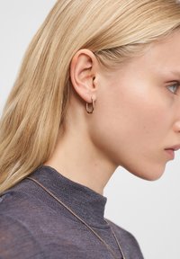 Blonde vrouw in een grijze hoge hals top, met een kleine, ronde gouden creool en een delicate gouden ketting, die naar rechts kijkt.