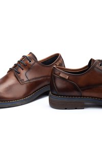 Zapatos de vestir de cuero marrón con un acabado suave, costuras en contraste, puntas redondeadas y diseño de cordones. Presentan acentos de logotipo en el costado.