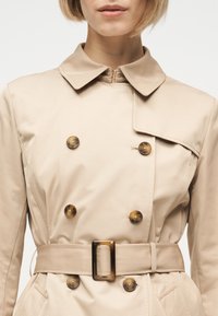 Γυναίκα που φορά μπεζ καμπαρντίνα διπλής ραφής με κουμπιά από κέλυφος χελώνας και ζώνη που ταιριάζει, σε απλό ανοιχτό φόντο.