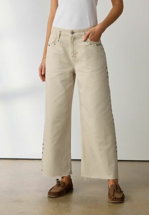 Person trägt beige Jeans mit weitem Bein und Metallnieten entlang der Taschen und Seiten, kombiniert mit braunen Mokassins auf einem glatten Boden.