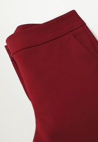 Pantalon sur mesure bordeaux avec une texture lisse, taille haute, fermeture éclair latérale et lignes épurées. Le tissu semble épais et structuré.