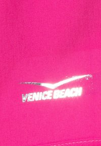 Vaaleanpunainen kangas, jossa on sileä pinta ja hopeanvärinen heijastava "VENICE BEACH" -logo sekä tyylitelty kuvio logon yläpuolella.